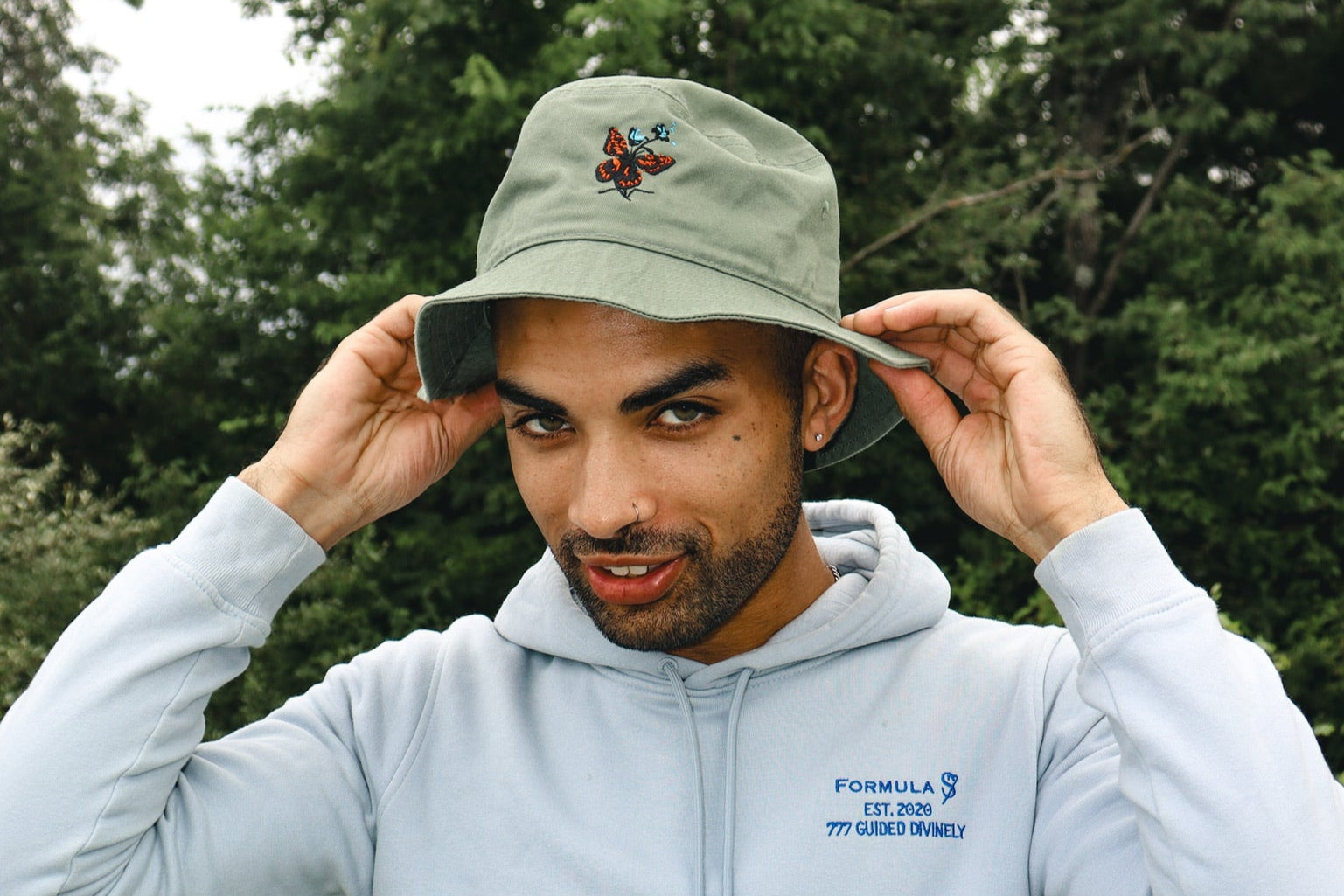 Man wearing a light blue hoodie with a 777 Angel number, adjusting a green cap with a butterfly outdoors.